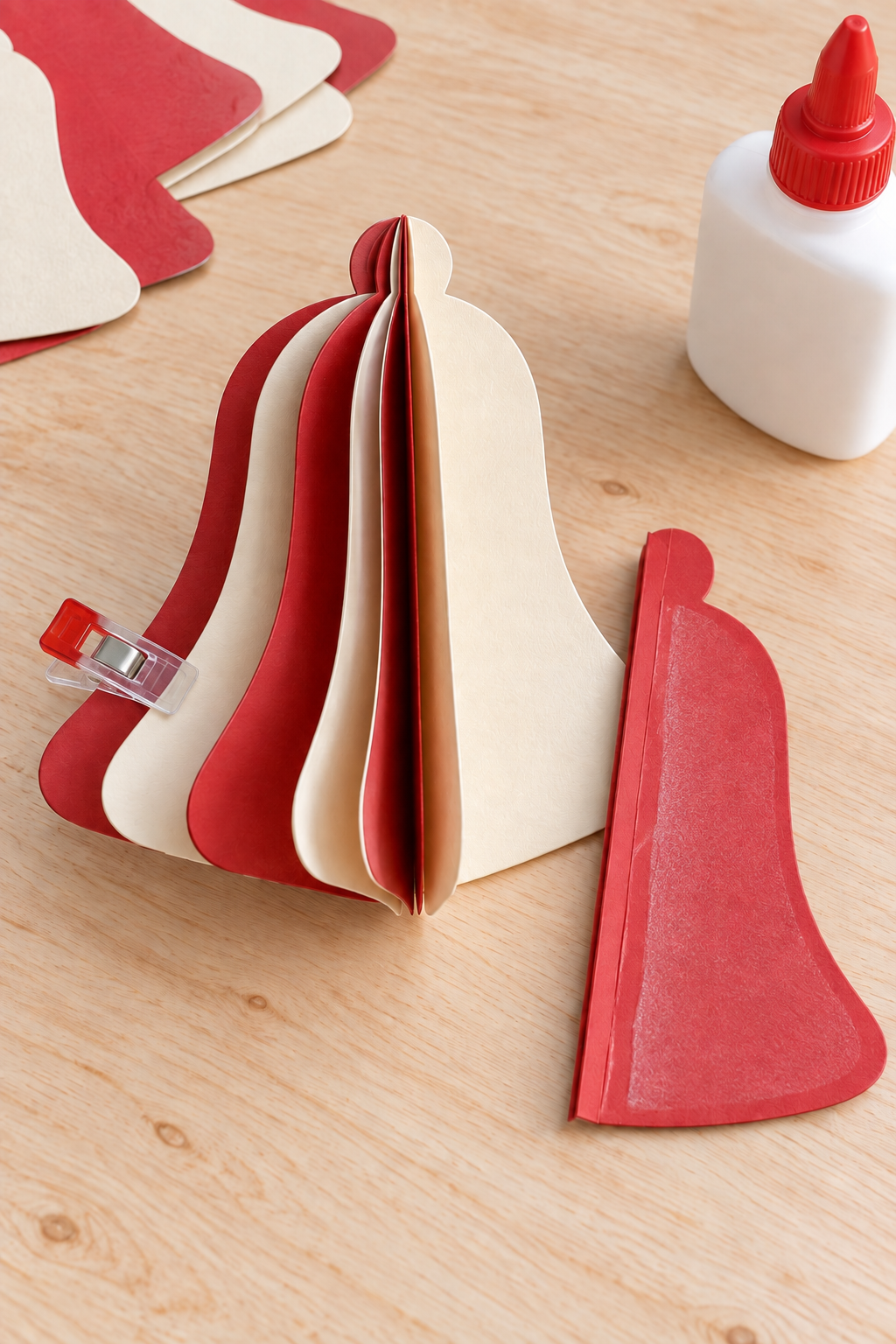 Red and cream folded bell pieces beside a glued red half and a small craft clip.