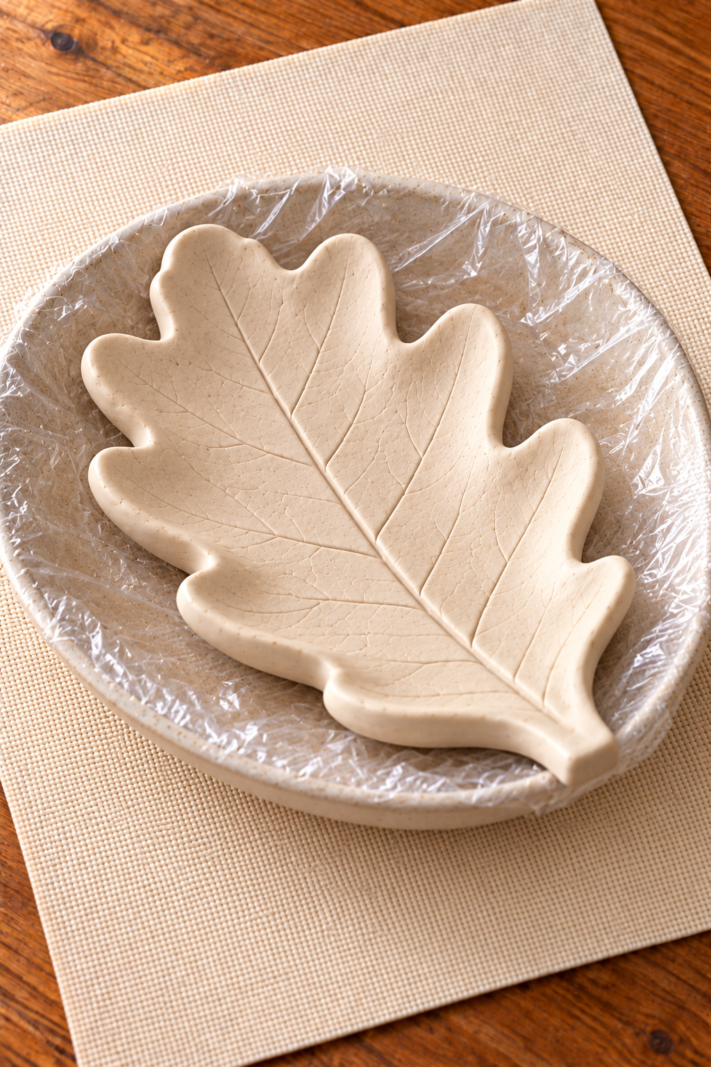 Unpainted leaf-shaped clay dish supported in a shallow saucer lined with plastic wrap.
