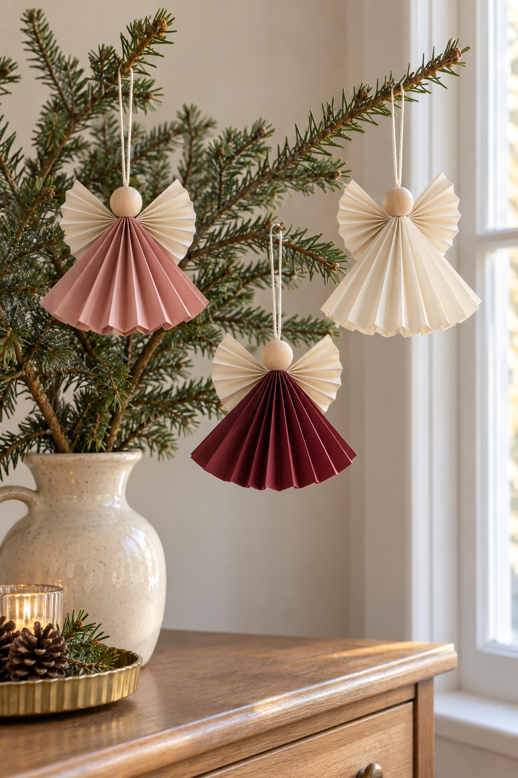 Rose, burgundy and cream paper angels with wooden heads hanging from evergreen branches.