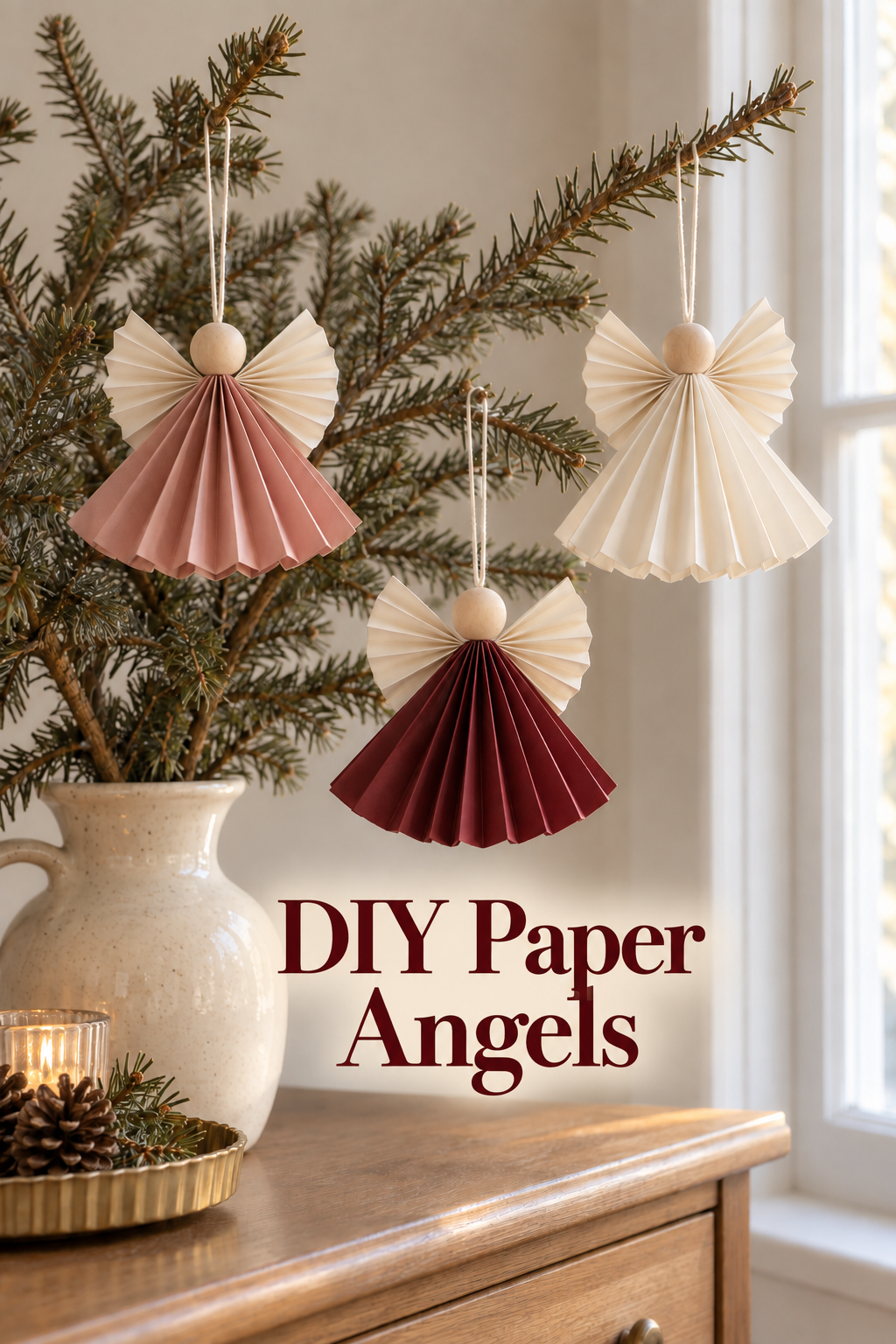 Rose, burgundy and cream paper angels with wooden heads hanging from evergreen branches.