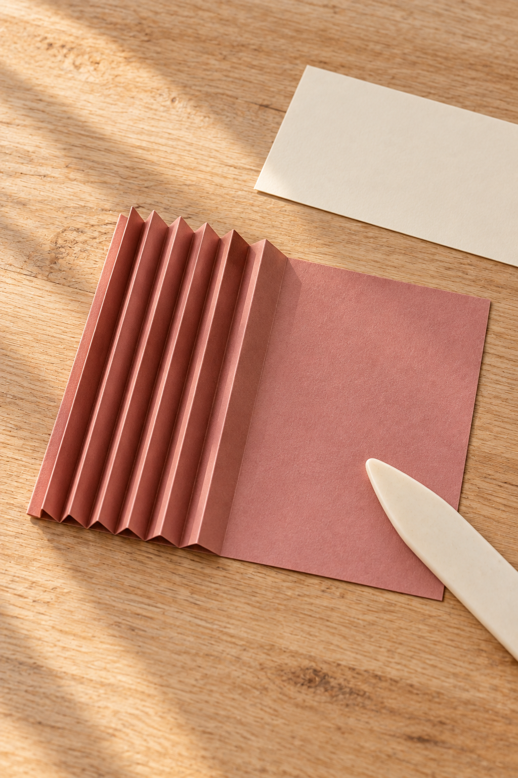 Dusty rose paper partly accordion folded beside a bone folder and a cream rectangle.