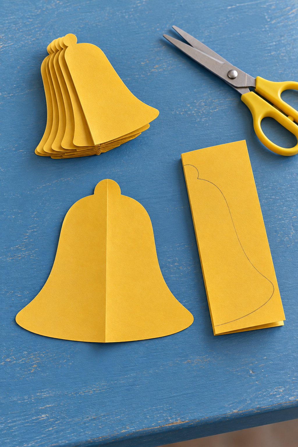 Yellow paper bell cutouts beside a traced folded guide and scissors on a blue table.