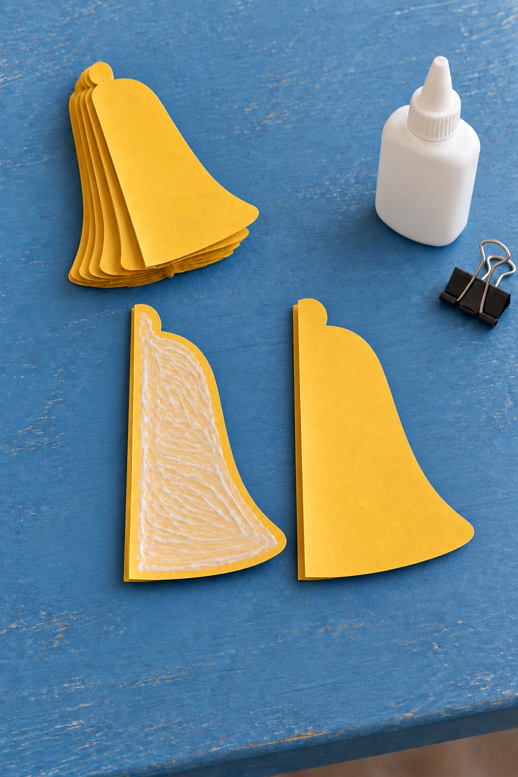 Glue coats one folded yellow half-bell beside matching cutouts, a bottle and binder clip.