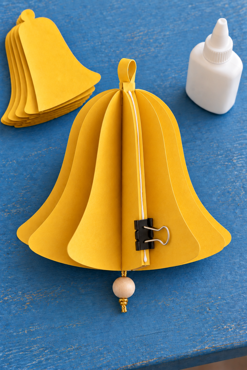 A binder clip holds the final glued seam of a fanned yellow paper bell with a bead below.
